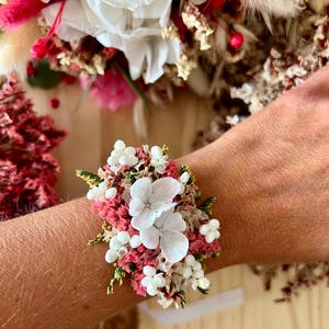 May include: A floral wrist corsage featuring dried white hydrangea petals, small white berries, pink statice, and green foliage. The arrangement is densely packed with preserved botanical elements, creating a textured, multi-colored cluster of natural dried flowers secured to a band.