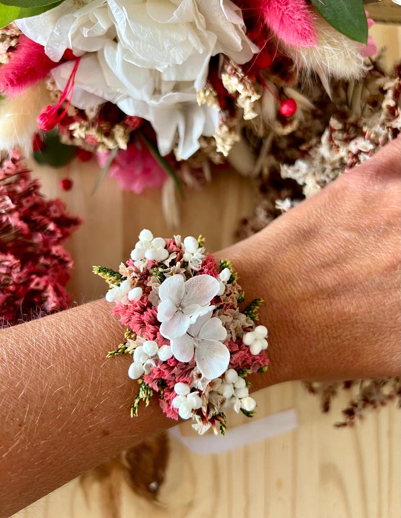 May include: A floral wrist corsage featuring dried white hydrangea petals, small white berries, pink statice, and green foliage. The arrangement is densely packed with preserved botanical elements, creating a textured, multi-colored cluster of natural dried flowers secured to a band.
