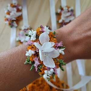 May include: A floral wrist corsage featuring white hydrangea petals, orange statice, small pink flowers, and green foliage. The arrangement is attached to a white satin ribbon, designed to be tied around the wrist for a decorative accessory.