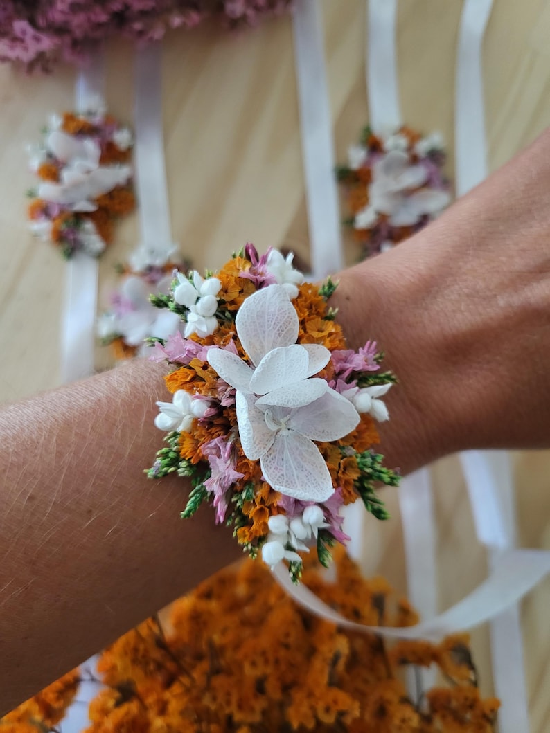 May include: A floral wrist corsage featuring white hydrangea petals, orange statice, small pink flowers, and green foliage. The arrangement is attached to a white satin ribbon, designed to be tied around the wrist for a decorative accessory.