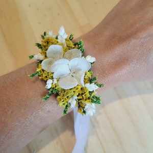 May include: A floral wrist corsage featuring white hydrangea petals, yellow statice, and green foliage accents. The arrangement is secured with a white ribbon tie, creating a layered botanical design that sits against the wrist with natural textures.
