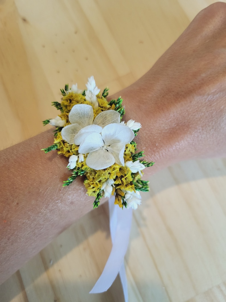 May include: A floral wrist corsage featuring white hydrangea petals, yellow statice, and green foliage accents. The arrangement is secured with a white ribbon tie, creating a layered botanical design that sits against the wrist with natural textures.