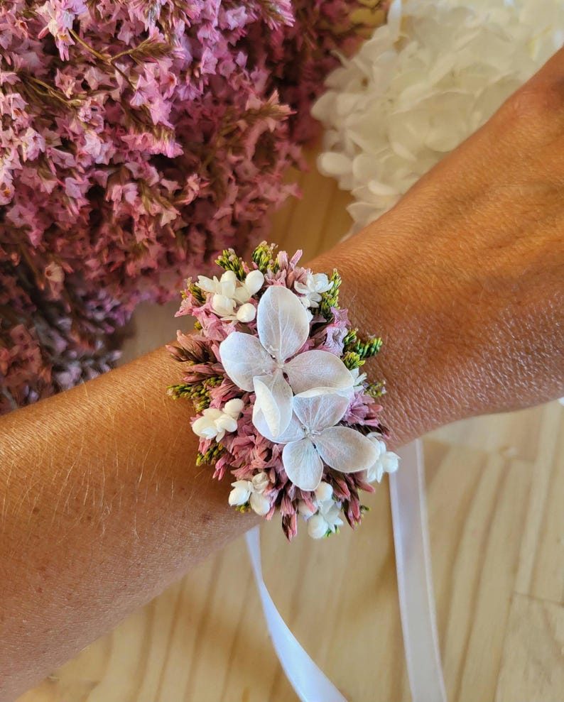 May include: A floral wrist corsage featuring dried pink statice, small white flowers, and two prominent white hydrangea petals. The arrangement is secured with a white ribbon tie, creating a textured botanical accessory with natural colors.