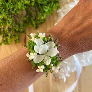 May include: A floral wrist corsage featuring white hydrangea petals, small white berries, and vibrant green foliage. The arrangement is secured with a white ribbon, creating a natural botanical accessory with a fresh, organic aesthetic.