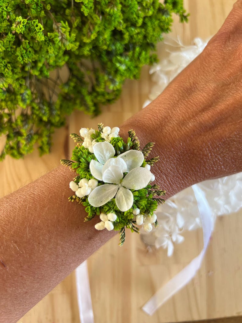 May include: A floral wrist corsage featuring white hydrangea petals, small white berries, and vibrant green foliage. The arrangement is secured with a white ribbon, creating a natural botanical accessory with a fresh, organic aesthetic.