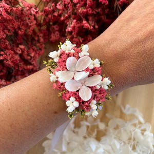 May include: A floral wrist corsage featuring a cluster of dried pink statice flowers, small white filler flowers, and two large, pale white hydrangea petals. The arrangement is secured with a white ribbon tied around the wrist.