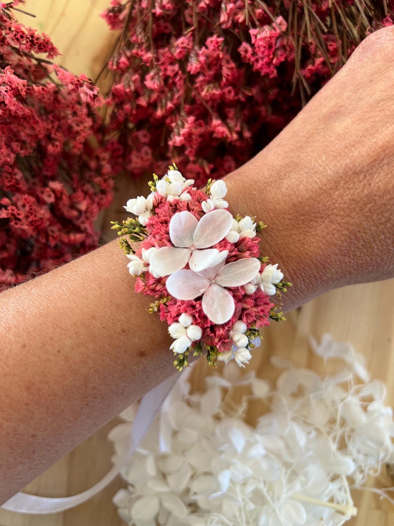 May include: A floral wrist corsage featuring a cluster of dried pink statice flowers, small white filler flowers, and two large, pale white hydrangea petals. The arrangement is secured with a white ribbon tied around the wrist.