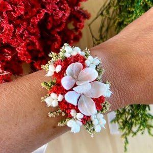 May include: A floral wrist corsage featuring a cluster of vibrant red statice flowers, small white blossoms, and delicate green foliage, centered with three large, pale white hydrangea petals arranged in a layered, circular pattern.
