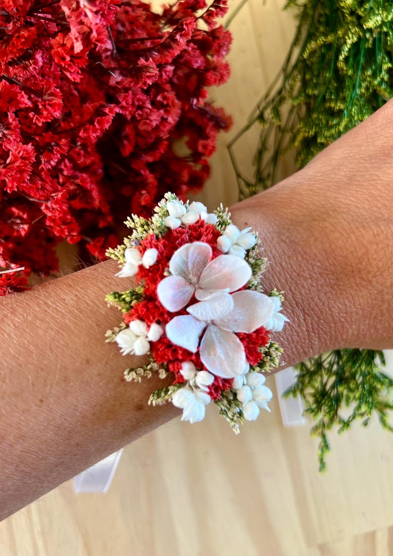 May include: A floral wrist corsage featuring a cluster of vibrant red statice flowers, small white blossoms, and delicate green foliage, centered with three large, pale white hydrangea petals arranged in a layered, circular pattern.
