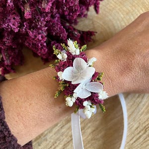 May include: A floral wrist corsage featuring deep magenta statice flowers, small white buds, and two delicate, translucent white hydrangea petals. The arrangement is secured to a thin, white satin ribbon for tying around the wrist.