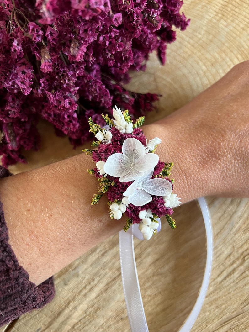 May include: A floral wrist corsage featuring deep magenta statice flowers, small white buds, and two delicate, translucent white hydrangea petals. The arrangement is secured to a thin, white satin ribbon for tying around the wrist.