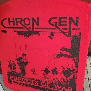 Op de afbeelding: Rood T-shirt met een zwarte grafische print van een band genaamd "Chron Gen" en de woorden "Puppets of War".