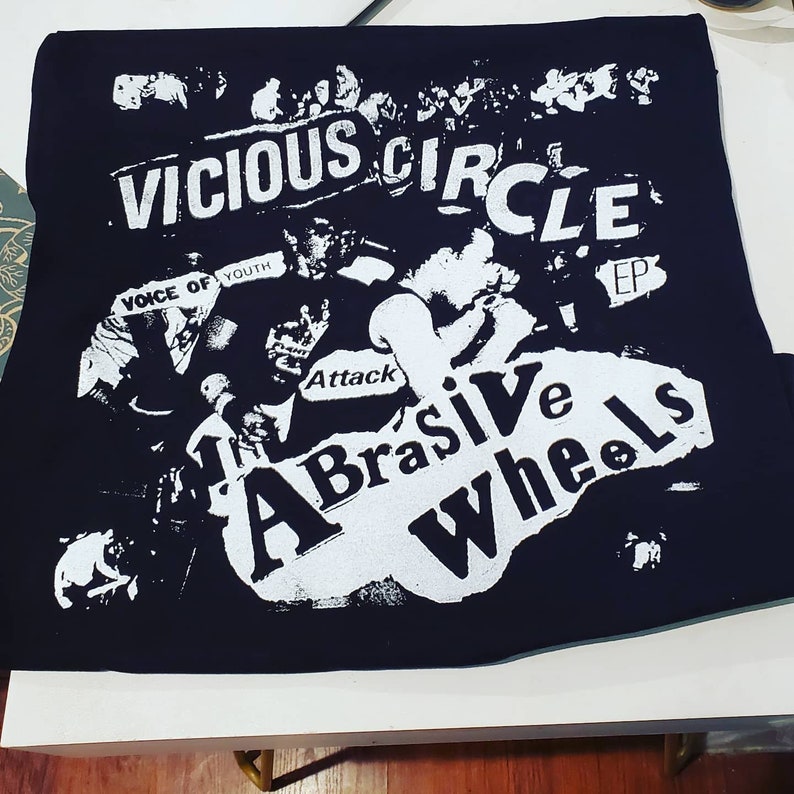 Pode incluir: Design gr&aacute;fico preto e branco em uma camiseta preta. O design apresenta o texto "Vicious Circle EP" e "The Abrasive Wheels" com um nome de banda "Voice of Youth" e "Attack" em texto menor.