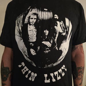 May include: A black short-sleeve t-shirt featuring a white graphic print of the band Thin Lizzy. The circular design displays three band members in a fisheye lens style, with the text THIN LIZZY printed in a bold, distressed font below the image.