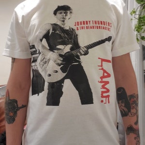 Puede incluir: Camiseta blanca con una imagen en blanco y negro de Johnny Thunders & The Heartbreakers. El nombre de la banda está impreso en rojo sobre la imagen. La palabra "L.A.M.F." está impresa en rojo en el lado derecho de la imagen.
