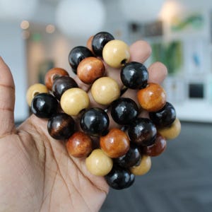 May include: A cluster of wooden beads in various colors, including black, light brown, medium brown, and beige. The beads are round and appear to be strung together. The beads are held in a hand.