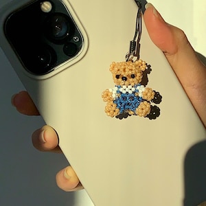 Könnte beinhalten: Beigefarbene Handyhülle mit einem Teddybär-Anhänger aus Perlen. Der Anhänger ist beige, blau und weiß, mit schwarzen Perlenaugen. Die Handyhülle hat eine Kameralinse und wird von einer Hand gehalten.