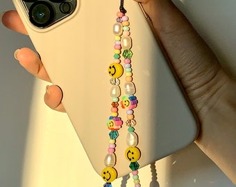以下が含まれることがあります： 白いパール、虹色のビーズ、黄色いスマイリーフェイスのチャームが付いたカラフルなビーズの携帯電話ストラップ。ストラップは白い携帯電話ケースに取り付けられています。
