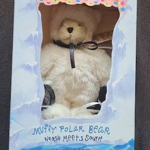 May include: White plush polar bear wearing a white hat and black ribbon. The bear is sitting in a box with a blue background and the words "Muffy Polar Bear North Meets South" printed on the front.