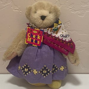 May include: A beige teddy bear wearing a purple dress with a crown pattern and a pink and white scarf with the text "Check Mates" on a red tag.