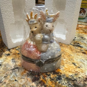 May include: A porcelain trinket box featuring a bear and a rabbit wearing crowns. The bear is wearing a red and white dress and the rabbit is wearing a blue and white dress. The box is decorated with stars and the year 2000 is written on the bear's crown.