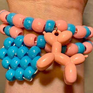 Pode incluir: Pulseira de contas rosa e azuis, com um berloque em forma de cão balão rosa. As contas redondas são enfiadas, criando um acessório colorido e original.