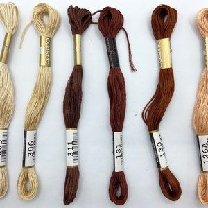 May include: Six spools of embroidery floss in various shades of brown, beige, and peach. The spools are labelled with the numbers 305, 306, 311, 131, 130, and 126A.