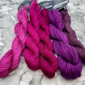 May include: Three skeins of embroidery floss in shades of pink, purple, and dark purple.