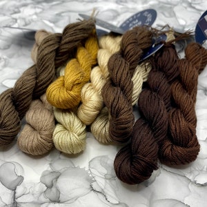 May include: A set of six skeins of embroidery floss in shades of brown, beige, and yellow. The floss is wound into small balls and arranged in a pile.
