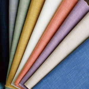 May include: A collection of folded linen napkins in various colors, including blue, navy, gray, green, yellow, white, peach, lavender, and beige. The napkins are arranged in a fan shape, showcasing their different hues and textures. The fabric appears to have a subtle woven pattern.
