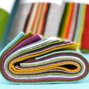 May include: A stack of colorful felt fabric sheets in various shades of green, blue, yellow, pink, purple, and gray. The felt is rolled up and the edges are visible.