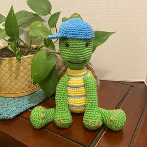 May include: A crocheted green turtle wearing a blue hat with yellow and blue stripes on its shell. The turtle is sitting on a wooden surface.