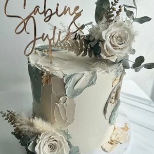 May include: A white wedding cake with gold accents and a brushstroke design. The cake is decorated with white flowers and greenery. The cake topper says "Sabine & Julian".
