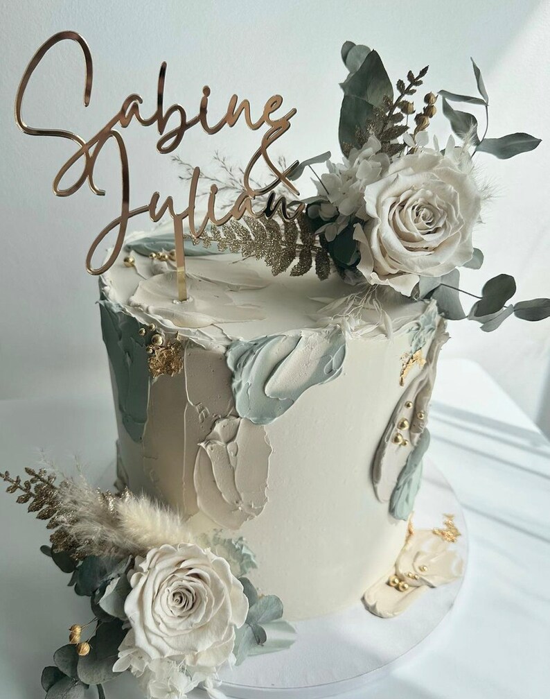 May include: A white wedding cake with gold accents and a brushstroke design. The cake is decorated with white flowers and greenery. The cake topper says "Sabine & Julian".