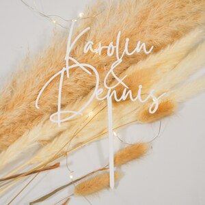 May include: White acrylic cake topper with the names "Karolin & Dennis" written in cursive script. The topper is placed on a background of dried pampas grass.