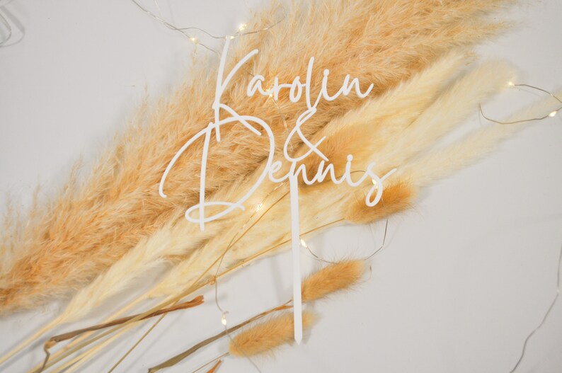 May include: White acrylic cake topper with the names "Karolin & Dennis" written in cursive script. The topper is placed on a background of dried pampas grass.