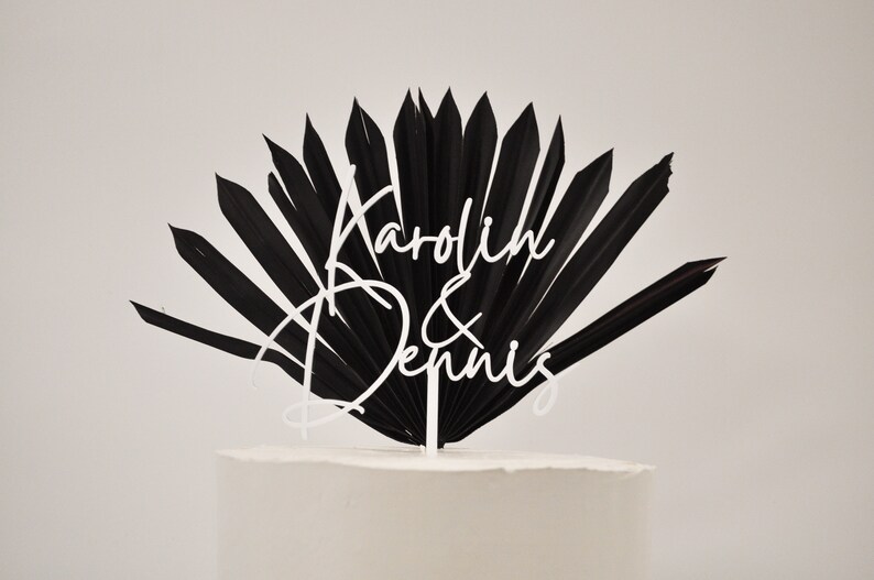 May include: Black palm leaf cake topper with white script lettering that reads "Karolin & Dennis".