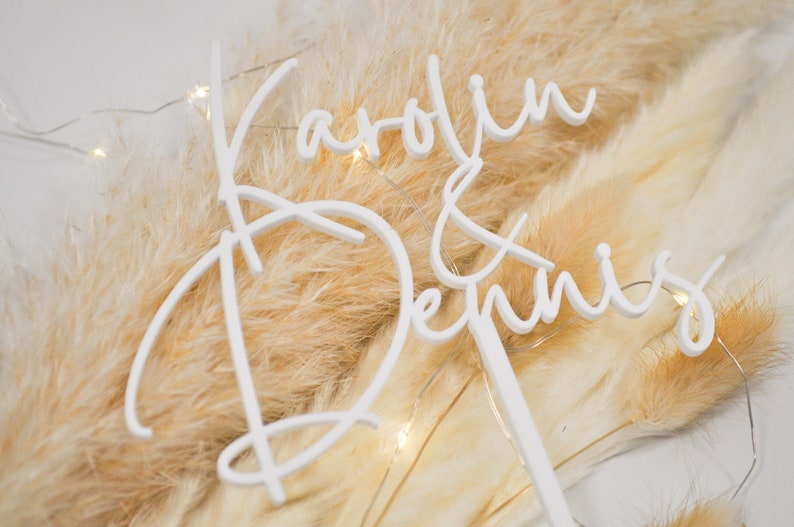 May include: White acrylic cake topper with the names "Carolyn & Dennis" written in a cursive script. The topper is placed on a bed of dried pampas grass.