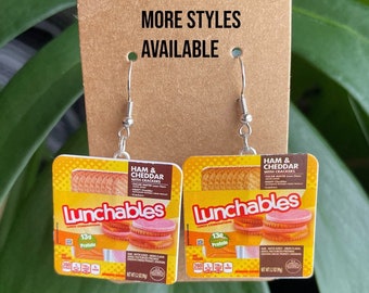 Może przedstawiać: Para kolczyków z miniaturowymi replikami opakowań przekąsek Lunchables Ham & Cheddar with Crackers. Kolczyki są srebrne z zapięciem na haczyk. Opakowanie Lunchables jest żółte z czerwonymi i białymi akcentami. Tekst na opakowaniu brzmi "Lunchables Lunch Combinations Ham & Cheddar with Crackers Oscar Mayer Lean Ham Kraft Cheddar Net Wt 2.3 oz (65g)"