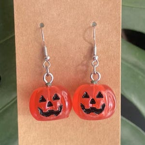 May include: A pair of orange pumpkin earrings with black carved faces, hanging from silver-colored hooks. The earrings are displayed on a brown card, with a green leaf in the background. These earrings are perfect for Halloween.