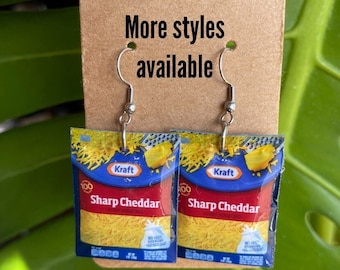May include: A pair of earrings featuring miniature Kraft Sharp Cheddar cheese packets. The earrings are silver and have a hook closure. The cheese packets are yellow and blue with the text "Kraft Sharp Cheddar" printed on them.