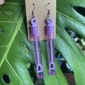 May include: A pair of purple bubble wand earrings. The wands are filled with clear liquid and have a silver hook.