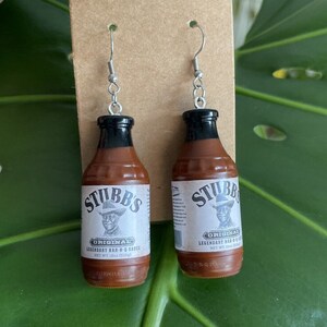 Op de afbeelding: Een paar zilveren oorbellen in de vorm van miniatuurflesjes Stubbs Original BBQ-saus. De flesjes zijn bruin met zwarte dopjes en hebben een wit etiket met het Stubbs-logo en de tekst: "Original Legendary Bar-B-Q Sauce Net Wt 18oz (510g)"