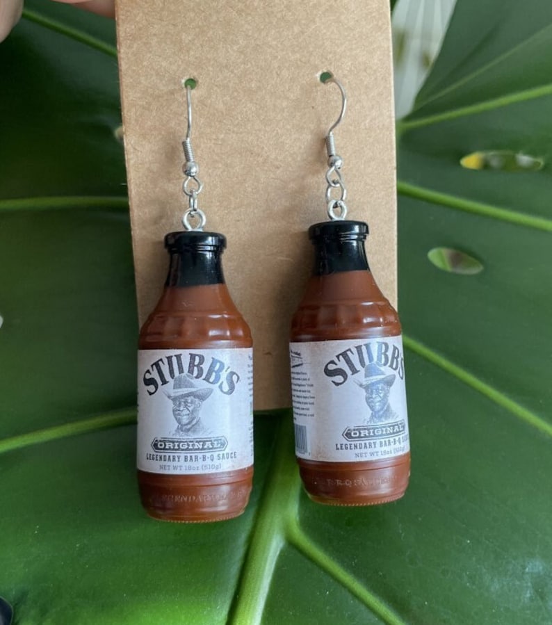 Op de afbeelding: Een paar zilveren oorbellen in de vorm van miniatuurflesjes Stubbs Original BBQ-saus. De flesjes zijn bruin met zwarte dopjes en hebben een wit etiket met het Stubbs-logo en de tekst: "Original Legendary Bar-B-Q Sauce Net Wt 18oz (510g)"