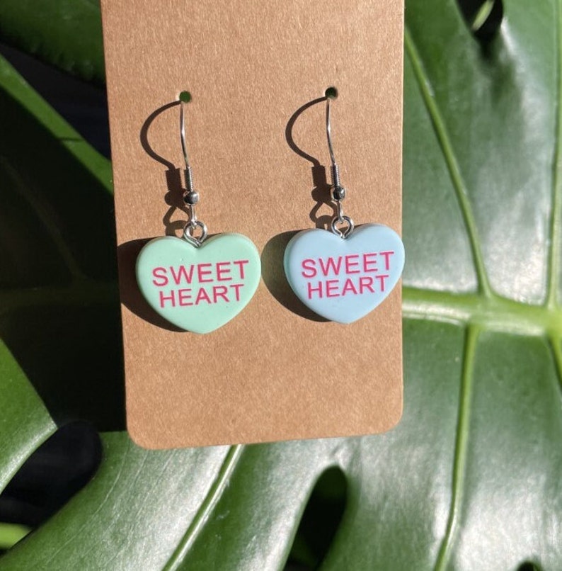 May include: A pair of silver heart-shaped earrings with the words "SWEET HEART" in pink lettering. One earring is light blue and the other is light green.