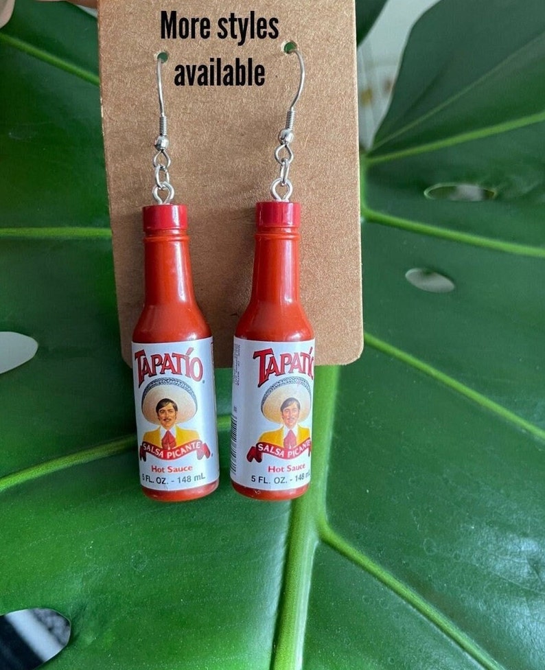 Op de afbeelding: Een paar rode hot saus fles oorbellen met zilveren haken. De flessen zijn gelabeld "TAPAT&Iacute;O SALSA PICANTE Hot Sauce 5 FL. OZ. - 148 mL."