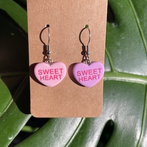 May include: A pair of pink heart-shaped earrings with the text "SWEET HEART" on each. The earrings are made of a pink and white plastic material and have silver hooks.