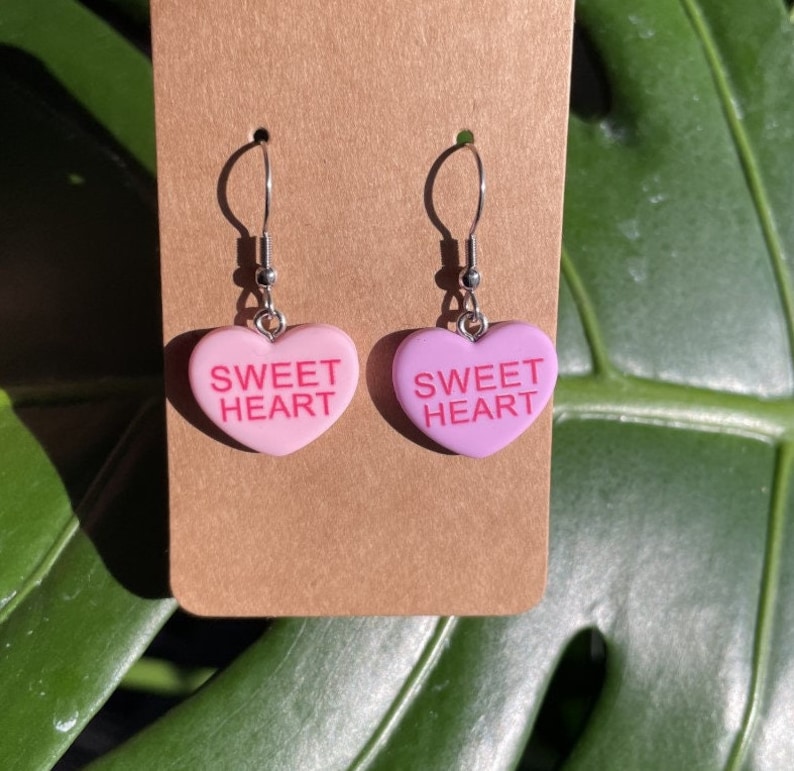 May include: A pair of pink heart-shaped earrings with the text "SWEET HEART" on each. The earrings are made of a pink and white plastic material and have silver hooks.