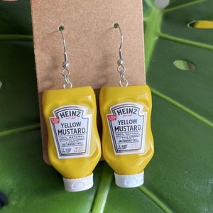 Op de afbeelding: Een paar gele mosterd fles oorbellen. De oorbellen zijn ontworpen om te lijken op Heinz Yellow Mustard flessen. De flessen hebben het Heinz logo en de woorden "Yellow Mustard" erop gedrukt.