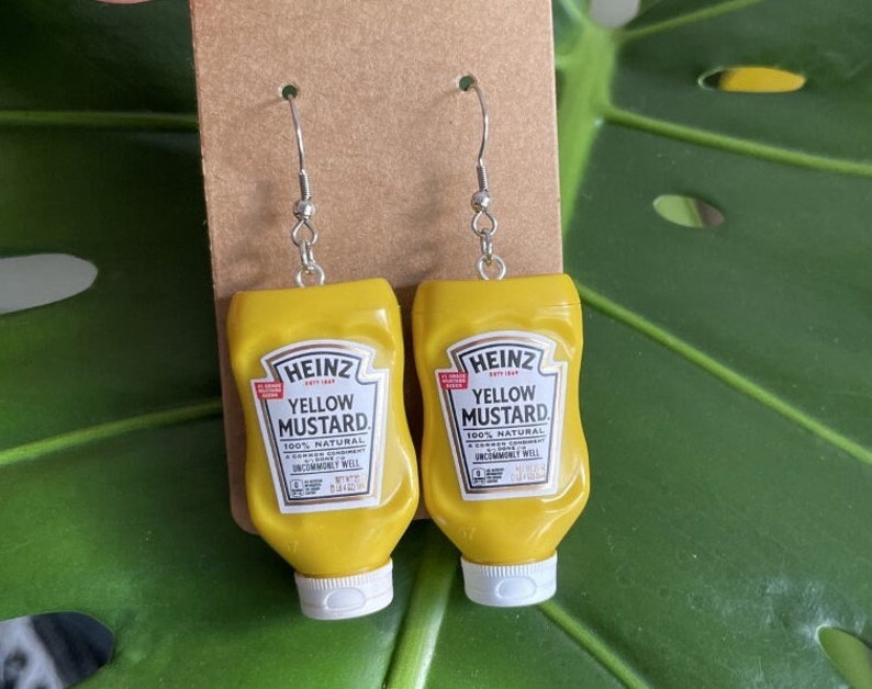 Op de afbeelding: Een paar gele mosterd fles oorbellen. De oorbellen zijn ontworpen om te lijken op Heinz Yellow Mustard flessen. De flessen hebben het Heinz logo en de woorden "Yellow Mustard" erop gedrukt.